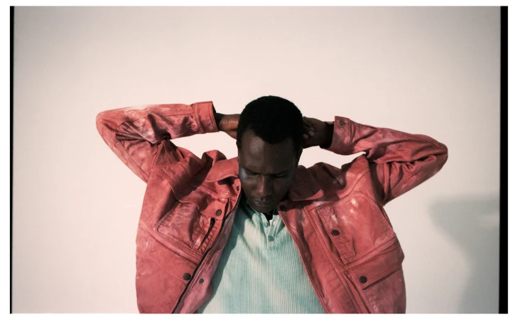 Man wearing a pink distressed leather jacket with arms raised behind his head against a white background