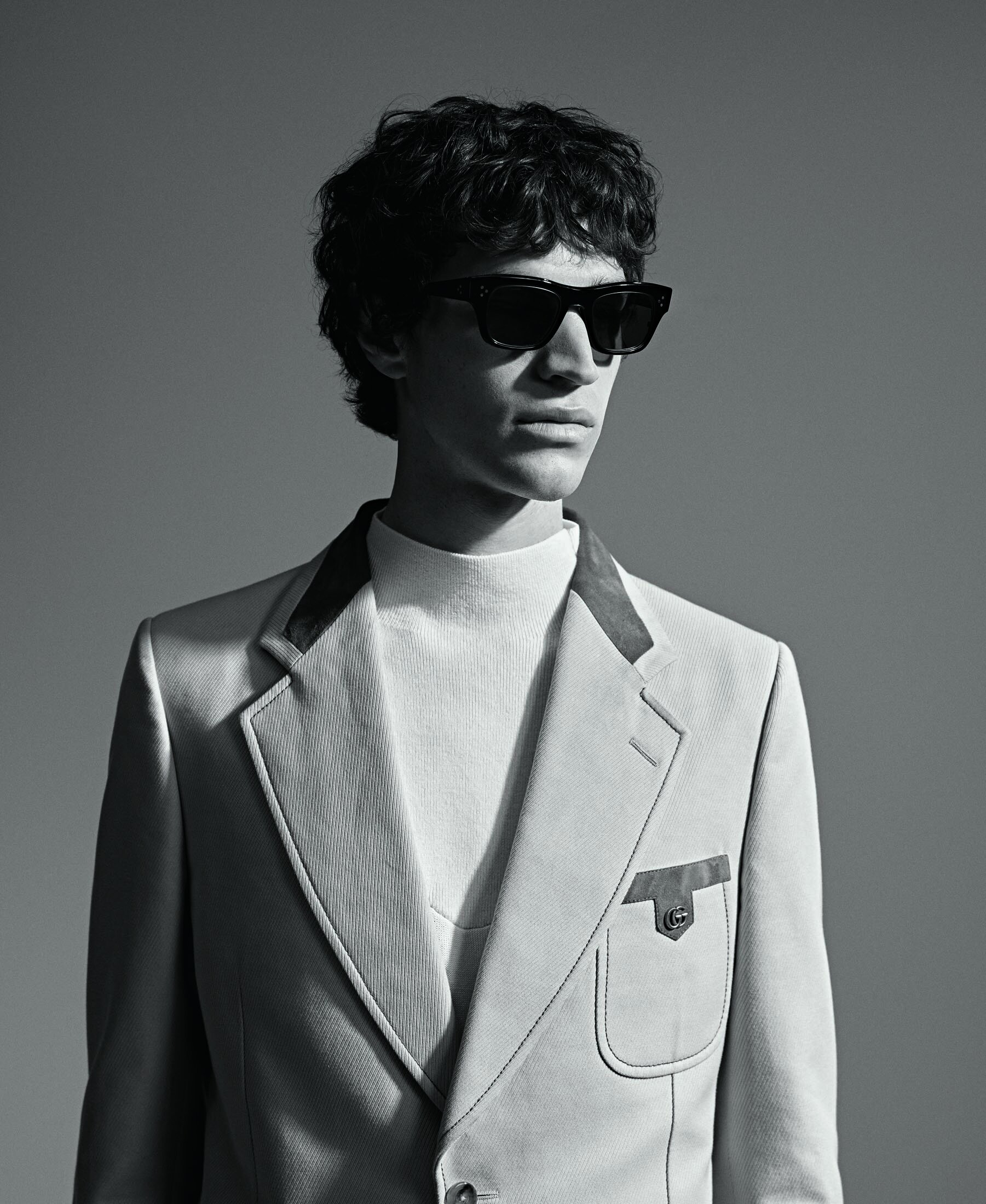 Monochrome portrait of a curly-haired man in sunglasses, a white turtleneck, and a light-colored blazer looking off-camera.