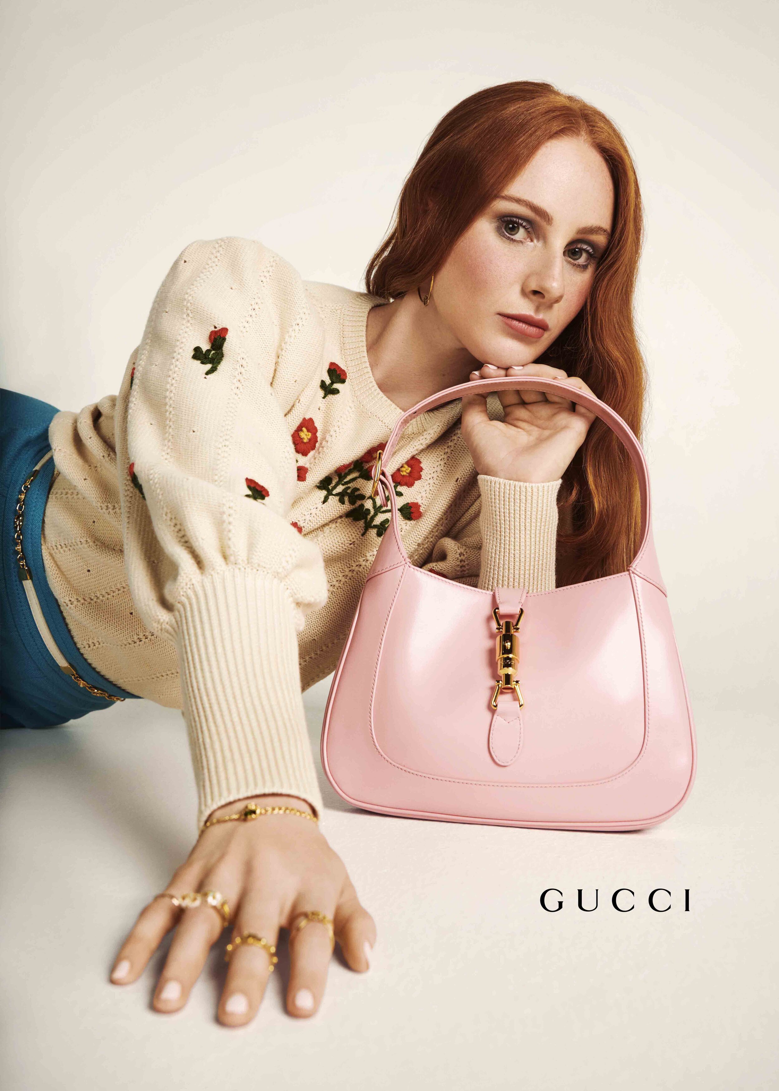 Fashion shot of a red-haired woman reclining with a pink Gucci purse, wearing a floral sweater and gold rings on a light background.