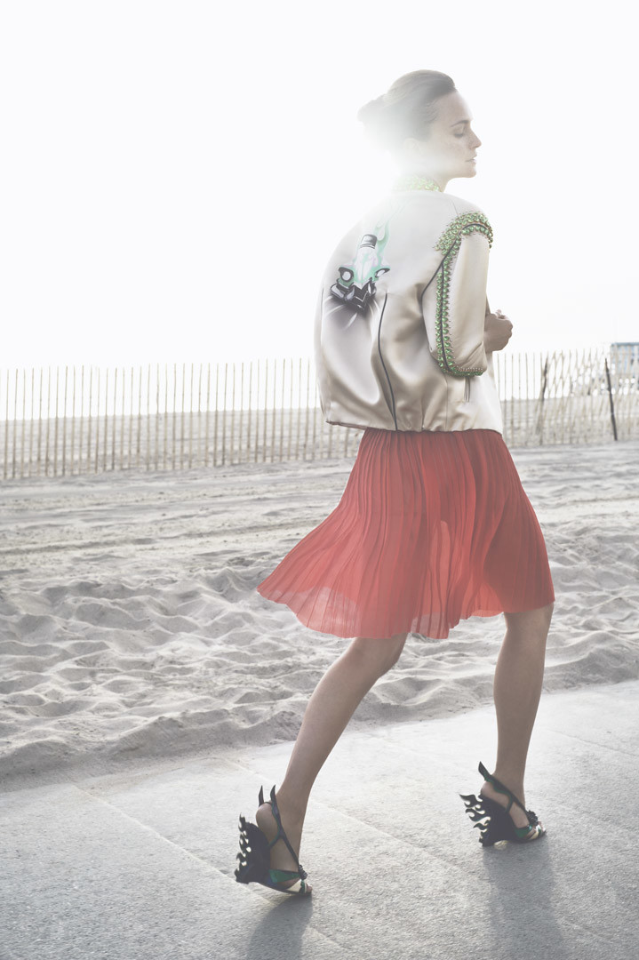 A woman in a peach jacket and red pleated skirt walks along a beach on a sunny day, fashion-forward footwear is visible.