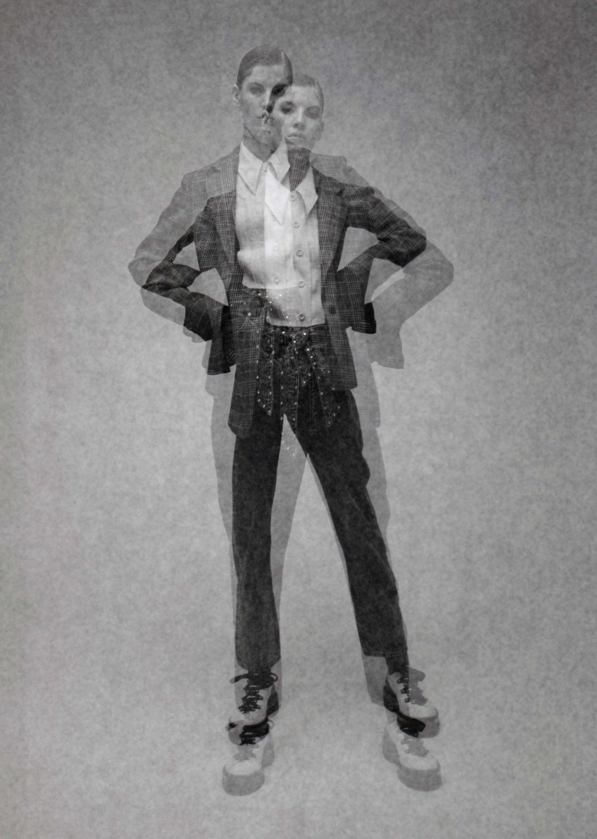Monochromatic portrait of a woman wearing a suit jacket and pants, posing with her hands on her hips against a faded backdrop.