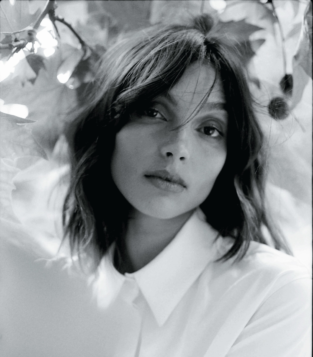 Monochrome portrait of a young woman with shoulder-length wavy hair, wearing a white collared shirt, against a blurred leafy background.