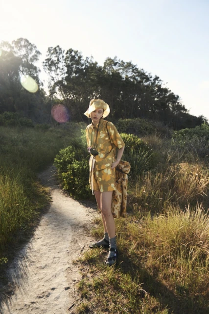 A person in a floral outfit stands on a grassy path with trees in the background, holding a camera, captured in natural light.
