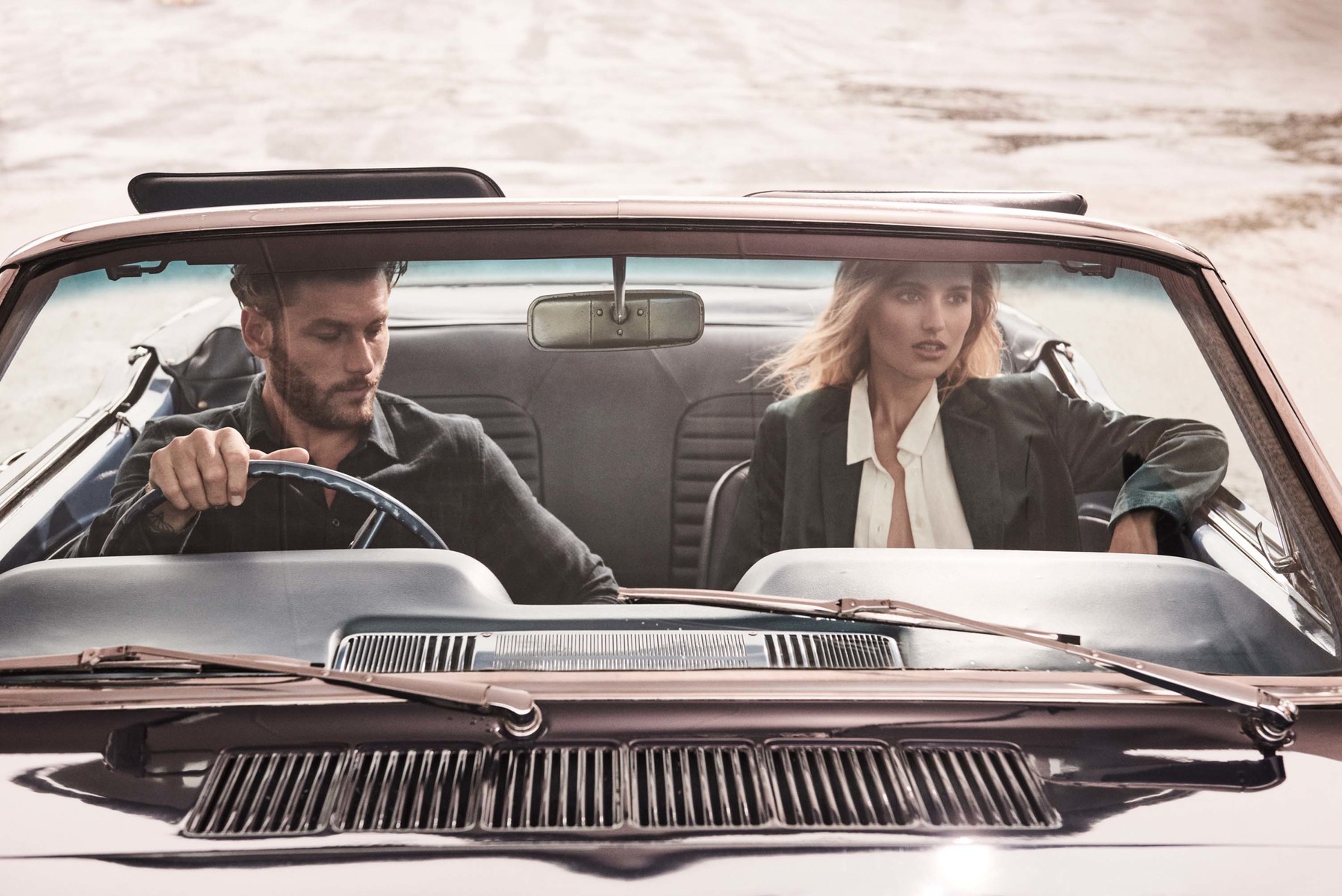 A man and a woman in a vintage convertible car on a sunny day, captured in a medium shot with a neutral color palette.
