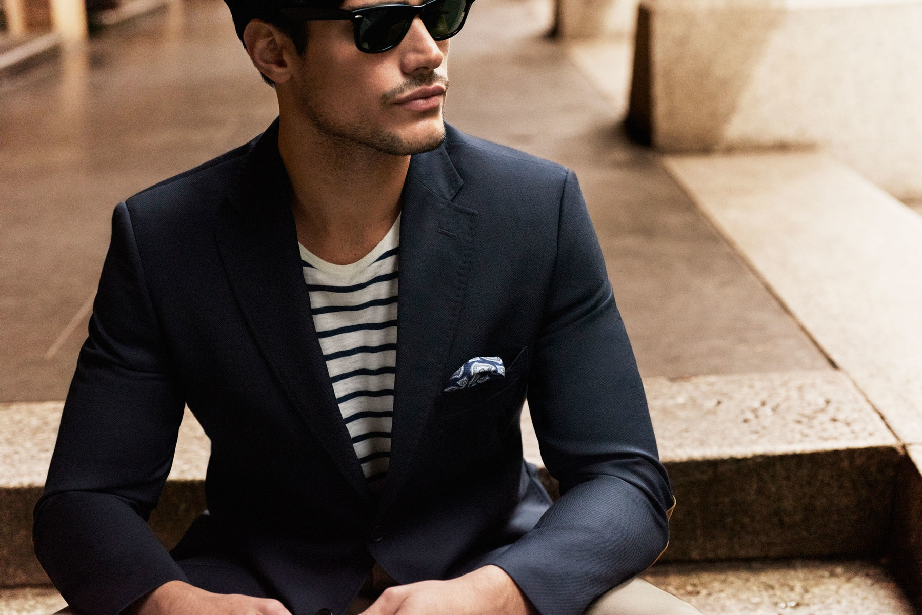 A stylish adult male in a dark blazer, striped shirt, and sunglasses poses outdoors. He is sitting on stone steps in neutral light.