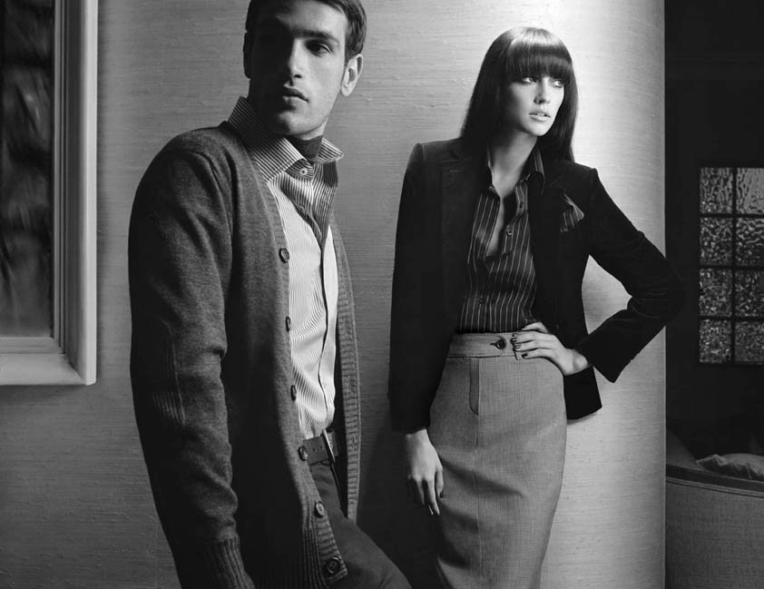 Fashion editorial shot of a male and female model in retro business attire in a staged indoor setting with monochrome color grade.