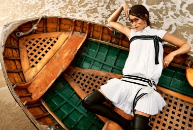 Woman in white pleated dress with black stripes and black boots reclining inside a wooden rowboat on sand