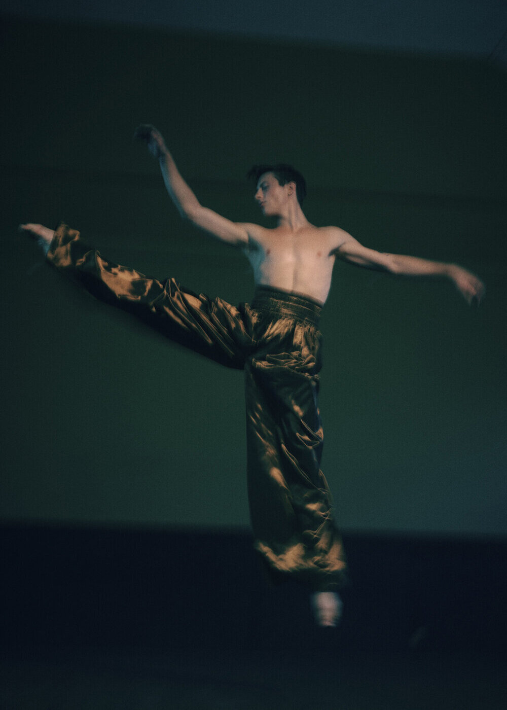 A shirtless adult male ballet dancer with brown hair is mid-air in a dance pose, wearing golden brown pants, on a green background.