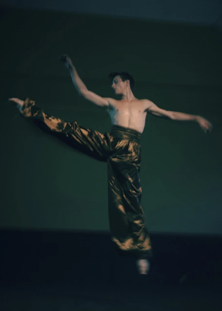 A shirtless adult male ballet dancer with brown hair is mid-air in a dance pose, wearing golden brown pants, on a green background.