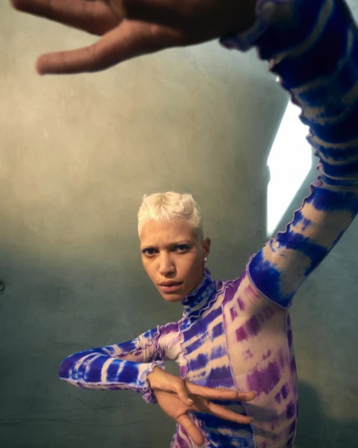 Woman with short blonde hair poses in a tie-dye long-sleeve top. The shot is at eye level, against a gray wall, with hands in frame.