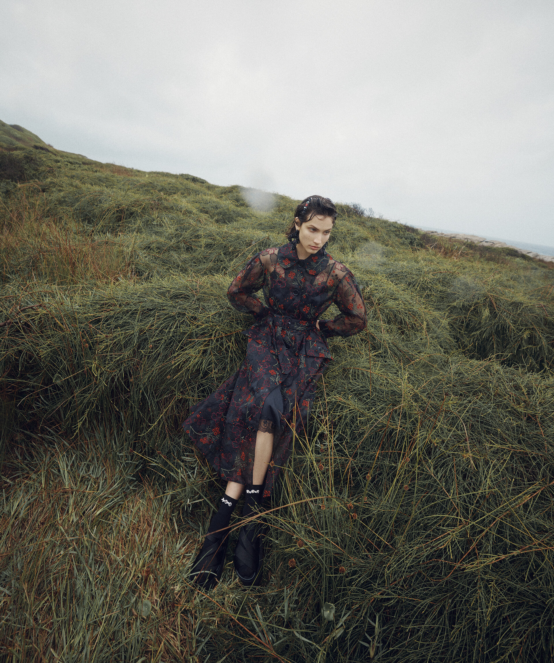 A model poses outdoors in a floral-patterned dress, black socks, and black boots, set against a grassy hillside and cloudy sky.