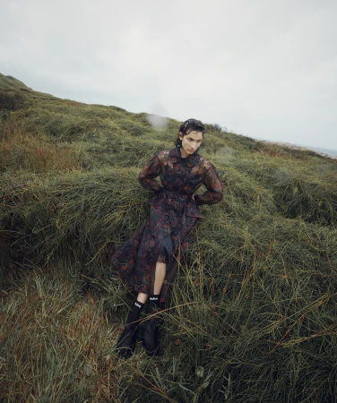 Model seated amongst tall green grass wearing a dark sheer patterned dress and black boots captured by Isaac Brown.