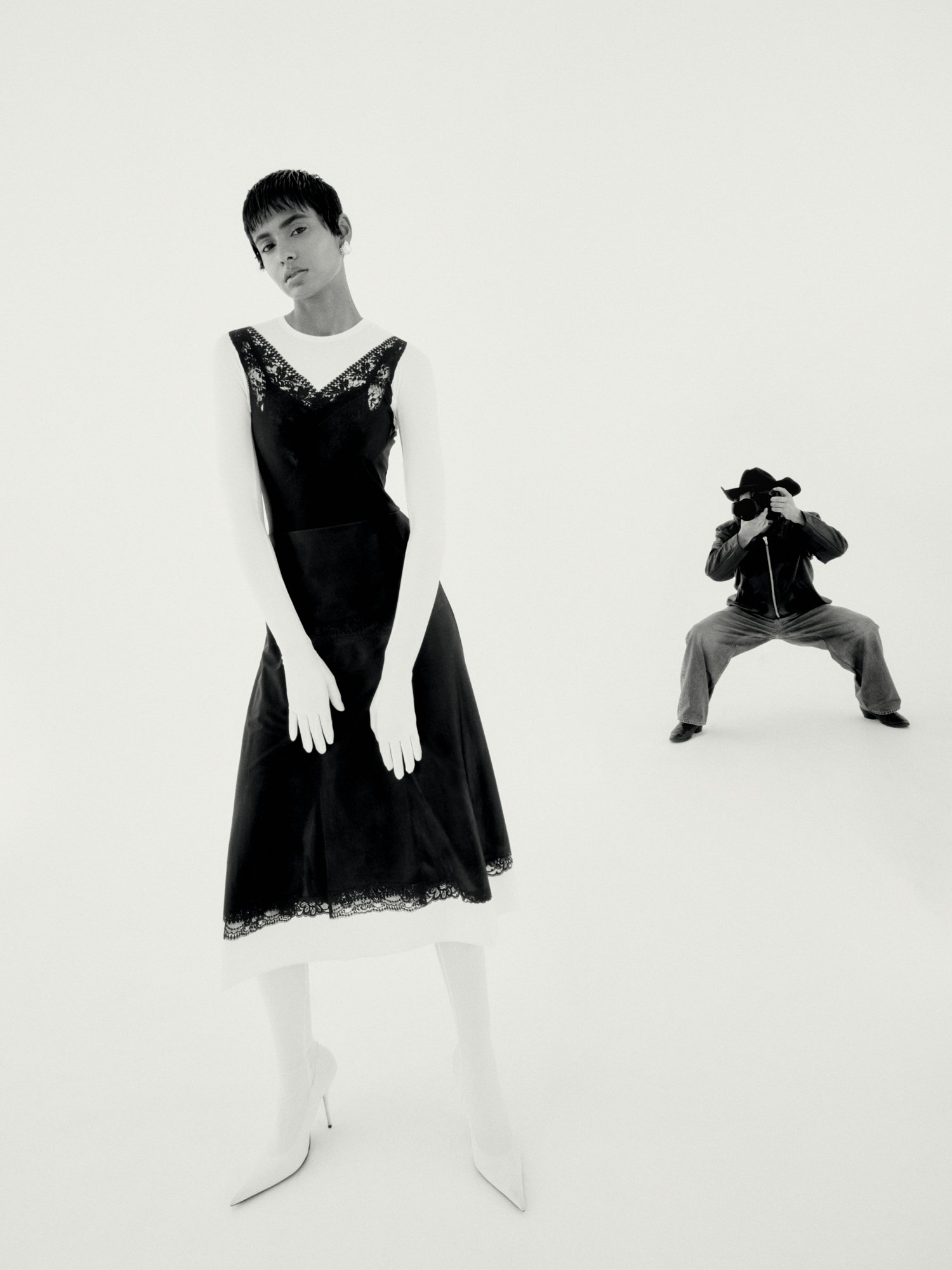 A fashion photograph shows a woman in a black dress and white gloves posing in a photo studio, with a photographer in the background.