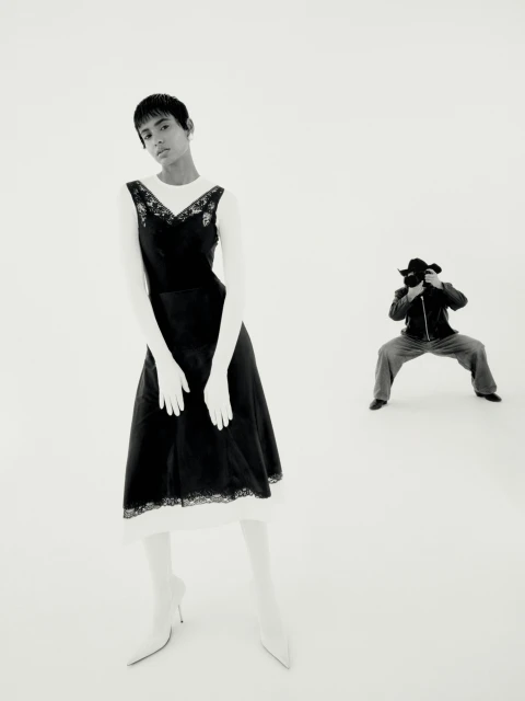 A fashion photograph shows a woman in a black dress and white gloves posing in a photo studio, with a photographer in the background.