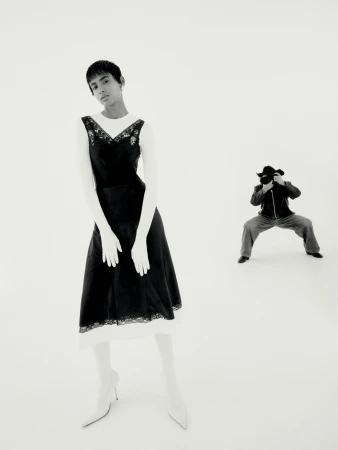 A fashion photograph shows a woman in a black dress and white gloves posing in a photo studio, with a photographer in the background.