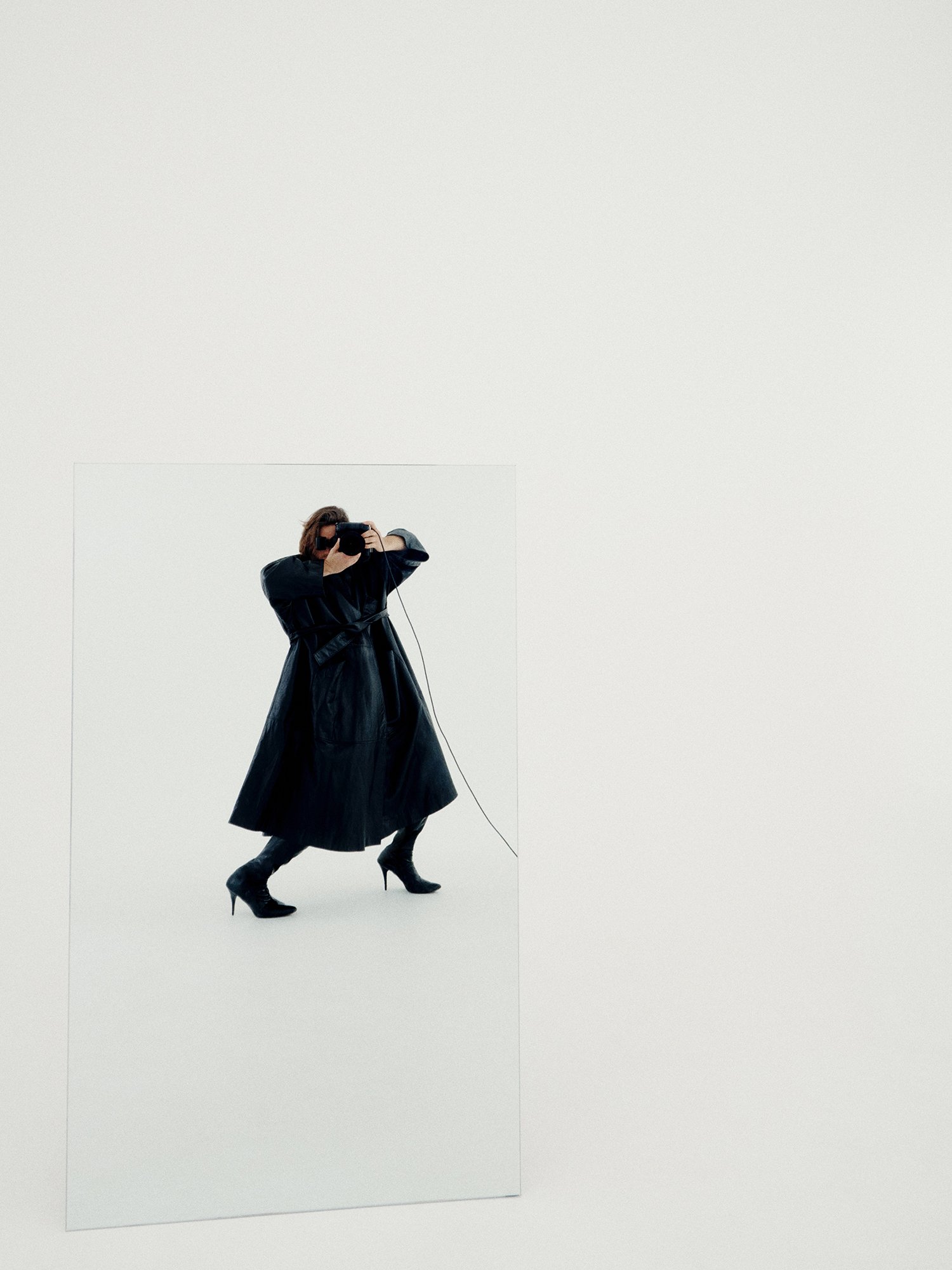 Woman in black coat and boots posing inside a mirror in a studio environment with a camera held to her face for a photoshoot.