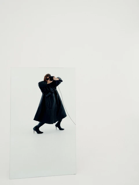 Woman in black coat and boots posing inside a mirror in a studio environment with a camera held to her face for a photoshoot.