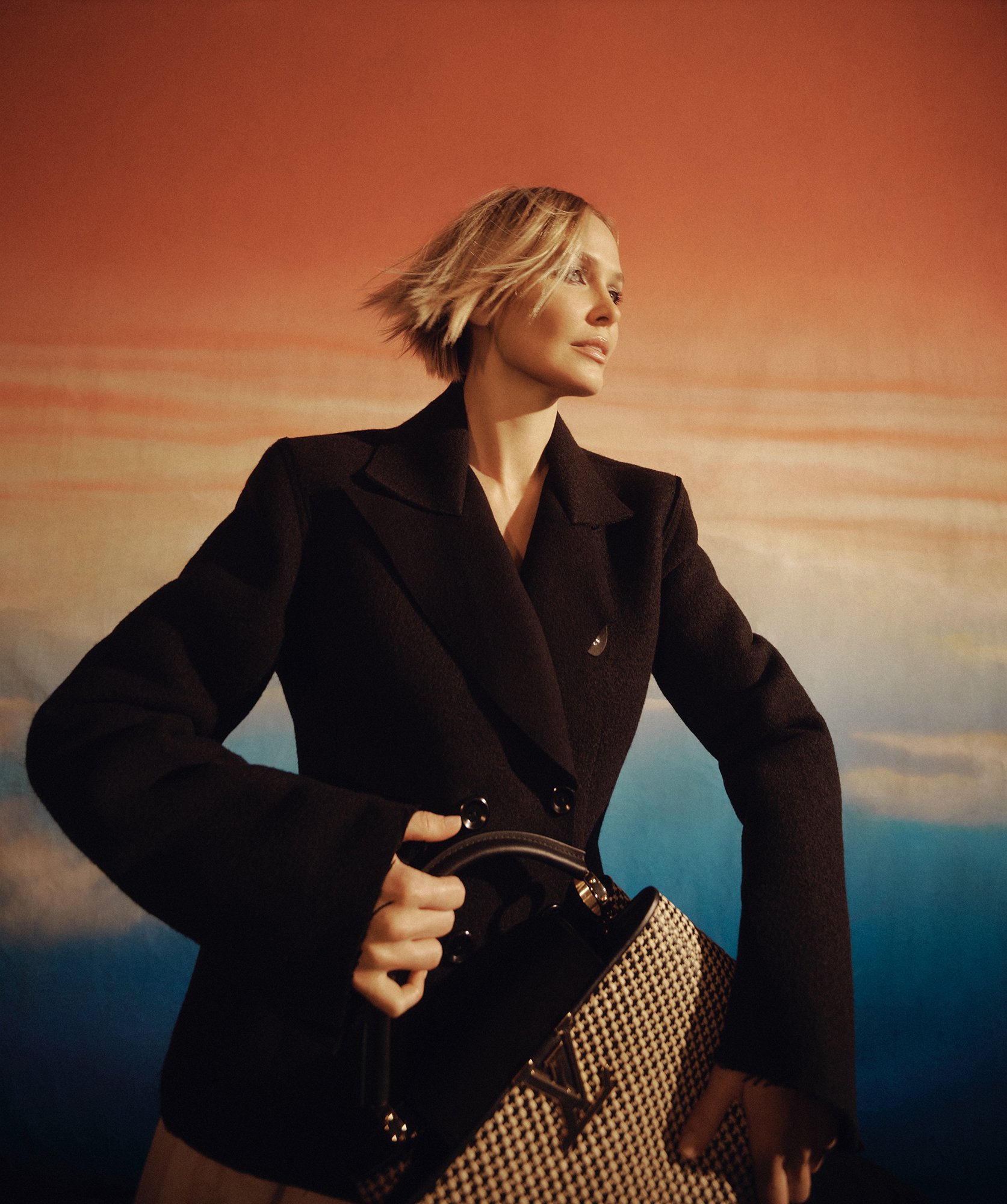 Fashion shot of a blonde woman in a black blazer holding a patterned bag, set against a gradient background with orange and blue tones.