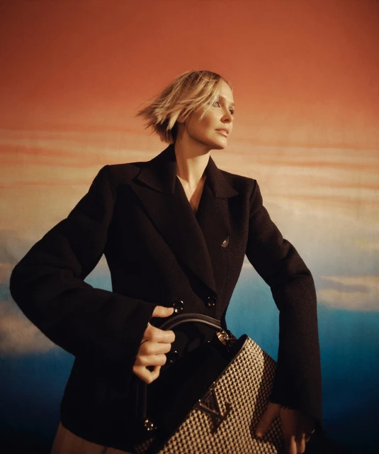 Fashion shot of a blonde woman in a black blazer holding a patterned bag, set against a gradient background with orange and blue tones.
