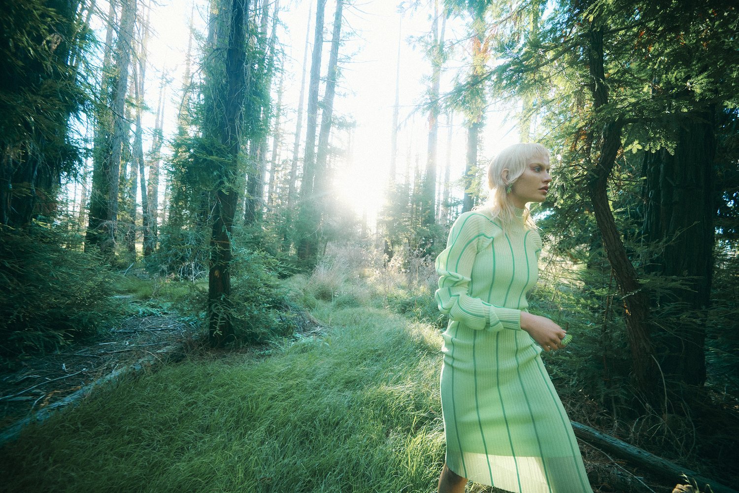 Woman in green dress walks through a forest with sun streaming through the trees, creating a bright, natural, outdoor environment.
