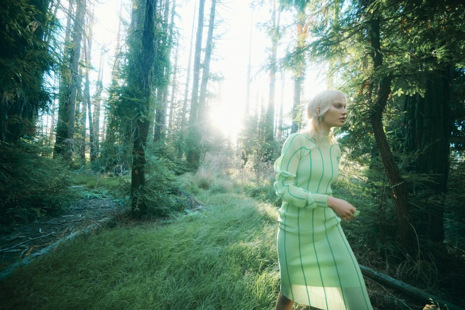 Woman in green dress walks through a forest with sun streaming through the trees, creating a bright, natural, outdoor environment.