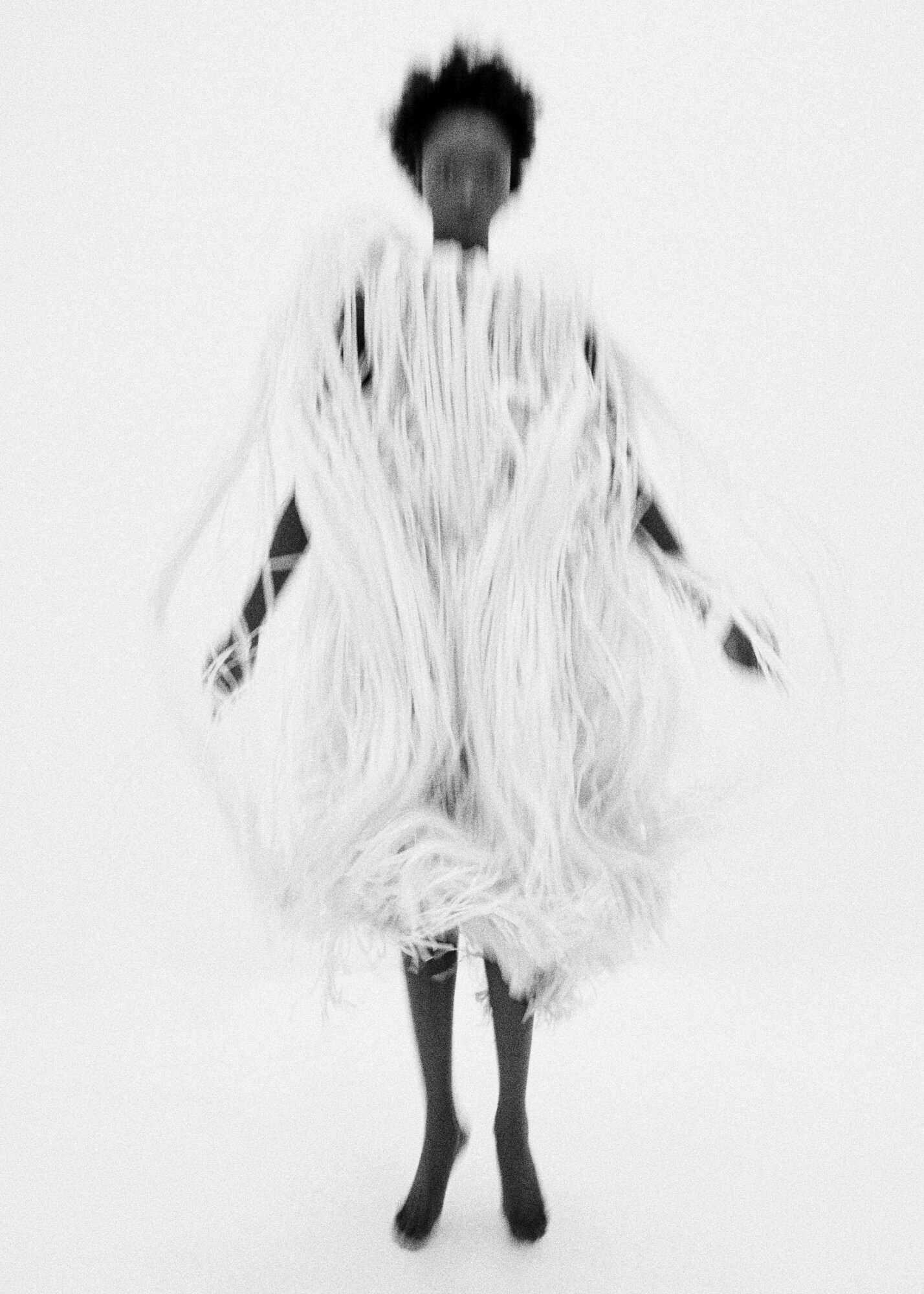 A single black woman stands in a studio wearing a full, long white dress of fringe, with arms out, against a stark white background.