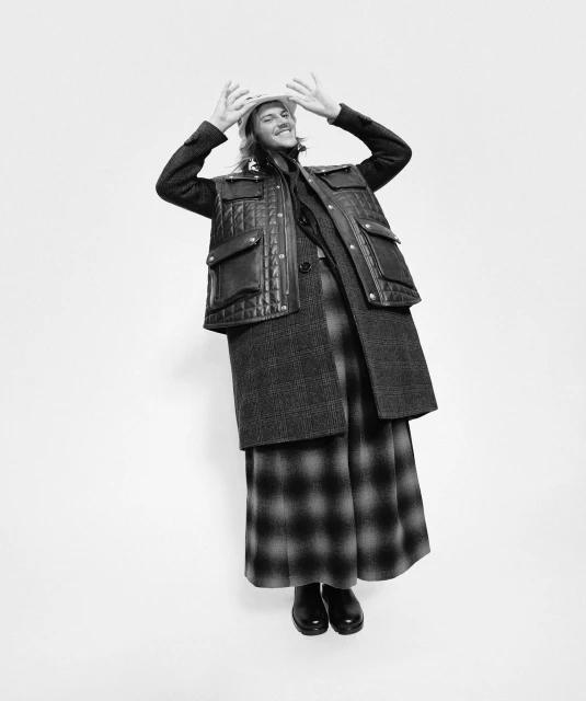 Full shot of a smiling adult in a wool coat, quilted vest, plaid skirt, boots and a brimmed hat, posing against a white backdrop.