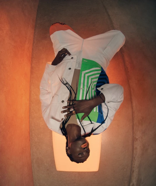 Adult wearing a white denim jacket with colorful accents and white pants posing in an orange-toned space with a light-up prop.