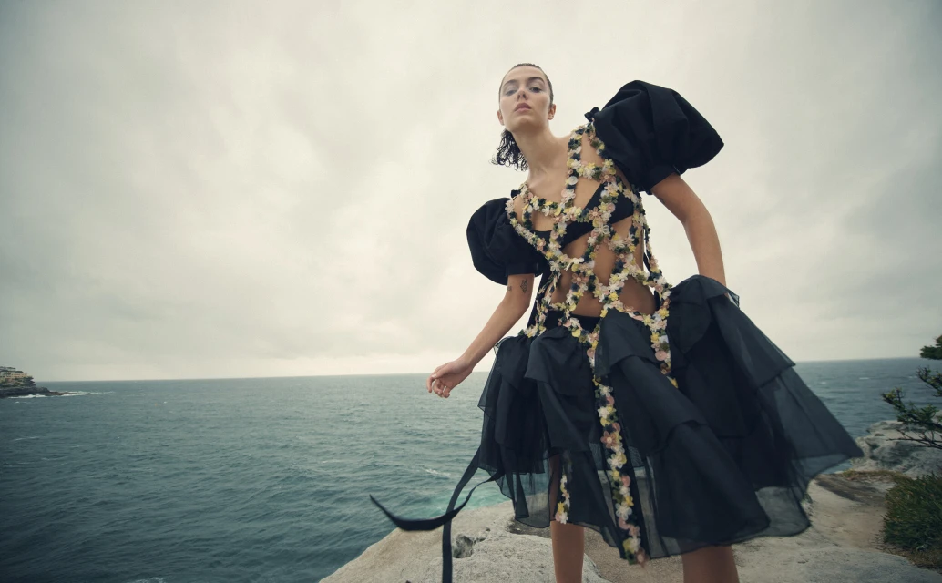 Fashion editorial shot of a model in a black, floral dress standing on a rocky coast with the ocean and cloudy sky in the background.