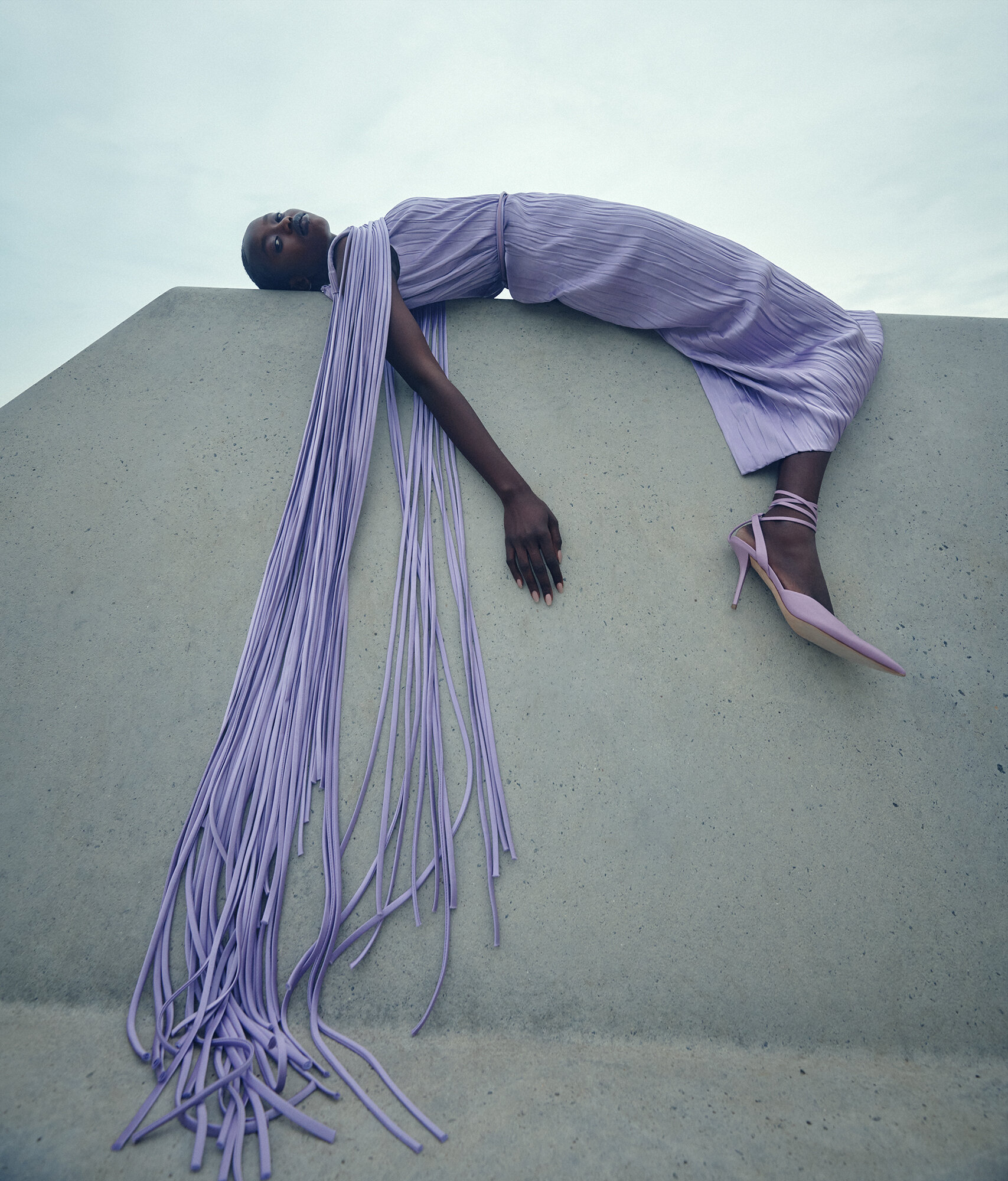 Fashion shot of a Black female model in a flowing lavender dress and matching heels, posing on a concrete structure against a sky background.
