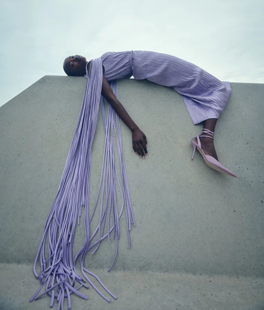 Fashion shot of a Black female model in a flowing lavender dress and matching heels, posing on a concrete structure against a sky background.