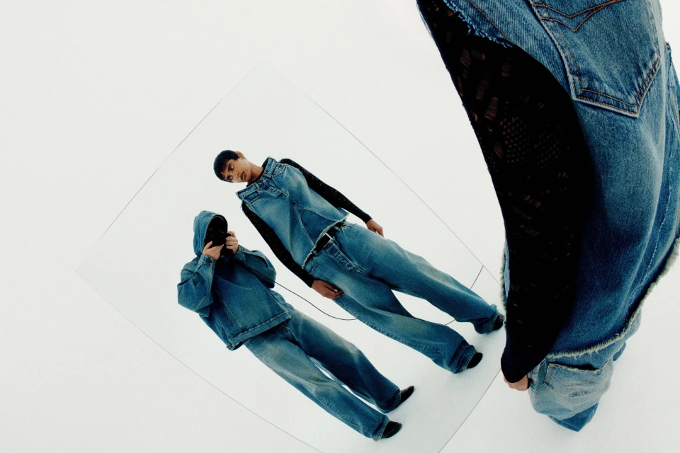 Two people in denim outfits are reflected in a mirror against a white background in a studio setting, showcasing fashion and style.