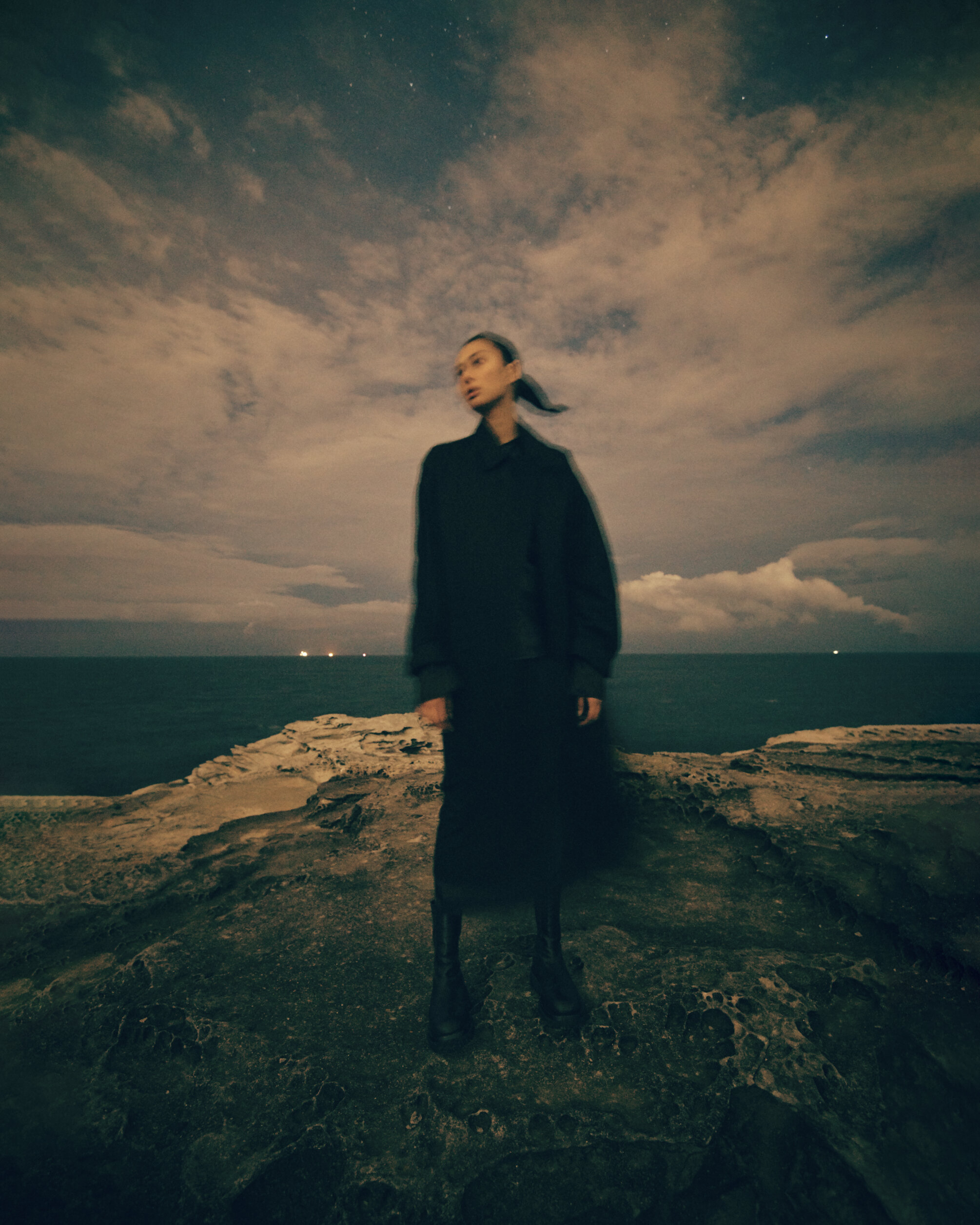 A blurry medium shot shows a woman in a dark dress standing on a rocky cliff at night with a cloudy sky and distant ocean view.