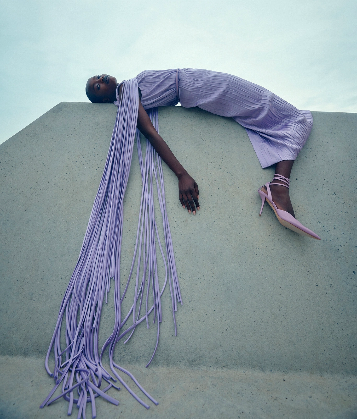 Model in lavender dress with long trailing fringe lying on a concrete structure against a pale sky.