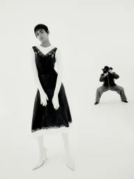 Model in a black slip dress standing in a white studio while a photographer crouches in the background