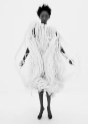 Model wearing a textured white fringed garment against a bright white background in a motion-blurred monochrome portrait
