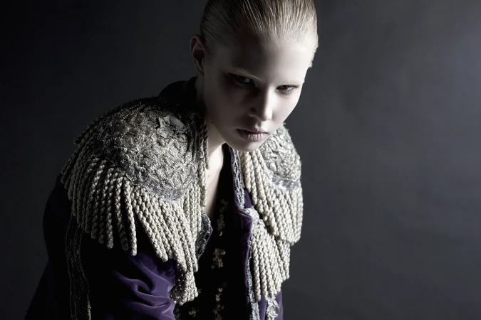 Portrait of a fair-skinned woman in a dark photo studio, wearing a purple velvet jacket with ornate silver shoulder embellishments.