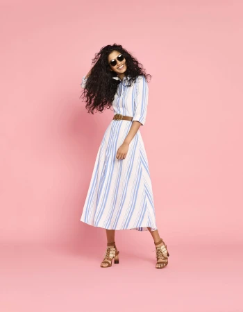 Smiling model with dark curly hair wearing a blue and white striped shirt dress and brown strapped heels against pink backdrop