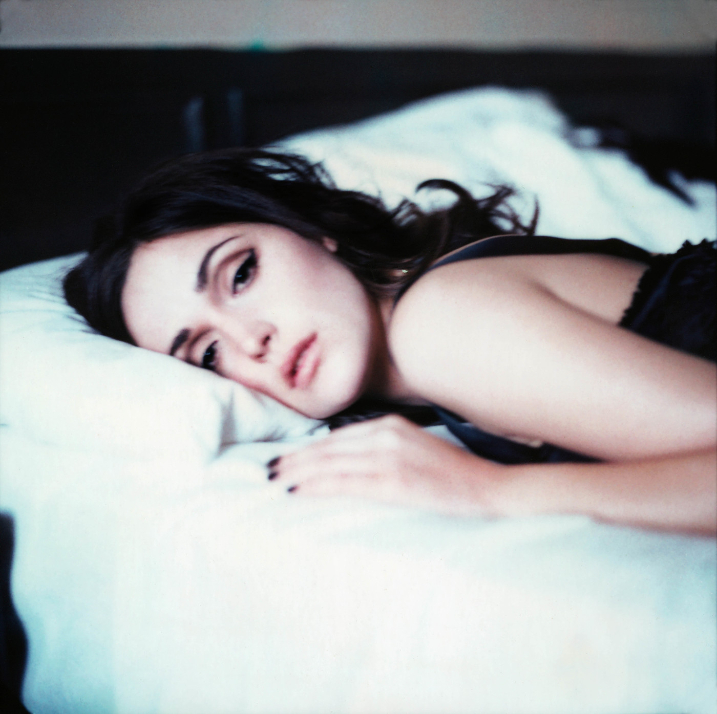 Woman lying on a white pillow in dim light. She has dark hair, is wearing black, and is gazing softly toward the camera.