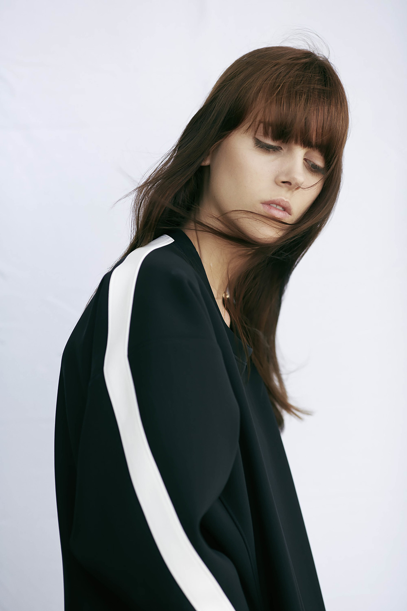 Brunette woman with bangs poses in a black top with a white stripe against a white backdrop in a studio portrait.
