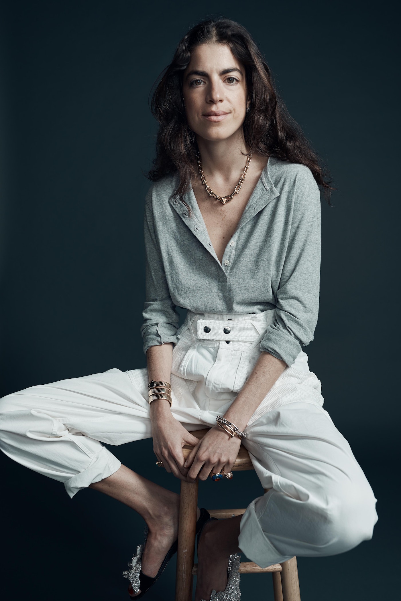 A brunette woman with a gold chain necklace, grey button-down shirt, and white pants sits on a wooden stool against a dark backdrop.