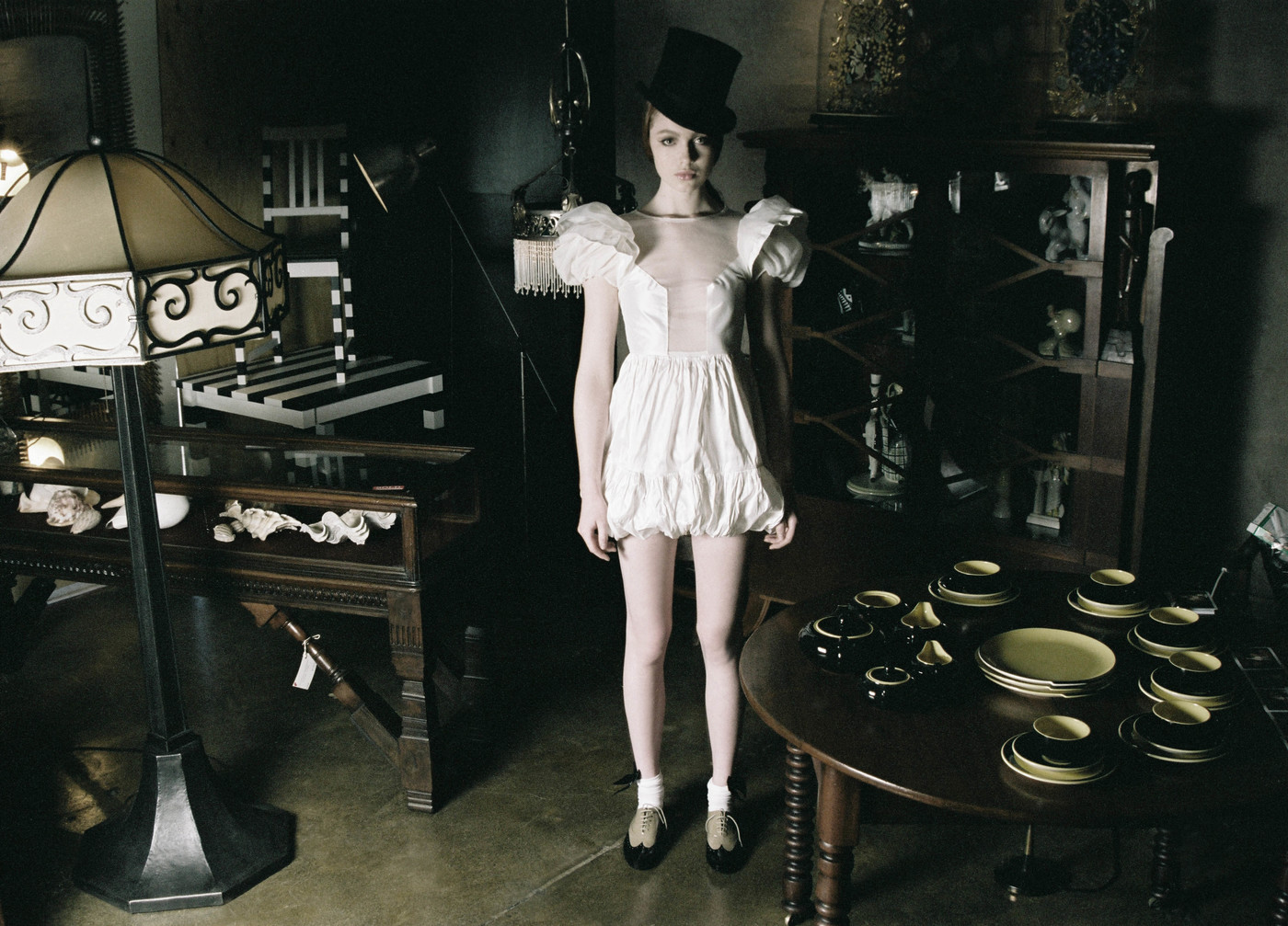 A slender young woman wearing a vintage white dress and top hat strikes a pose in an antique-filled room with moody lighting.
