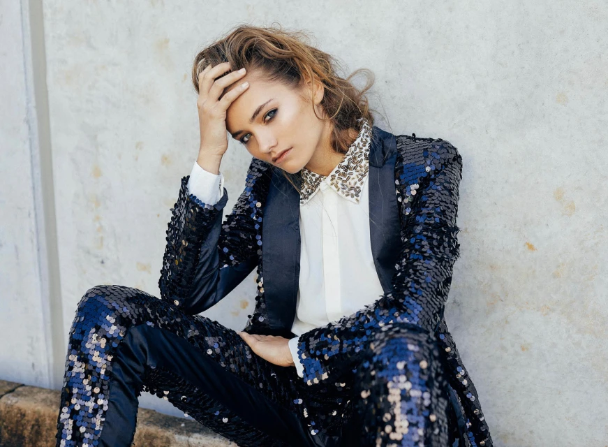 Woman poses wearing a sequined suit with a collared shirt in front of a textured wall.  Fashion portrait.  Eye-level angle.