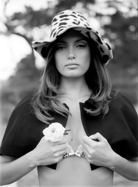 Black and white portrait of a woman with long hair and a patterned hat, holding a white rose, posing outdoors in soft light.