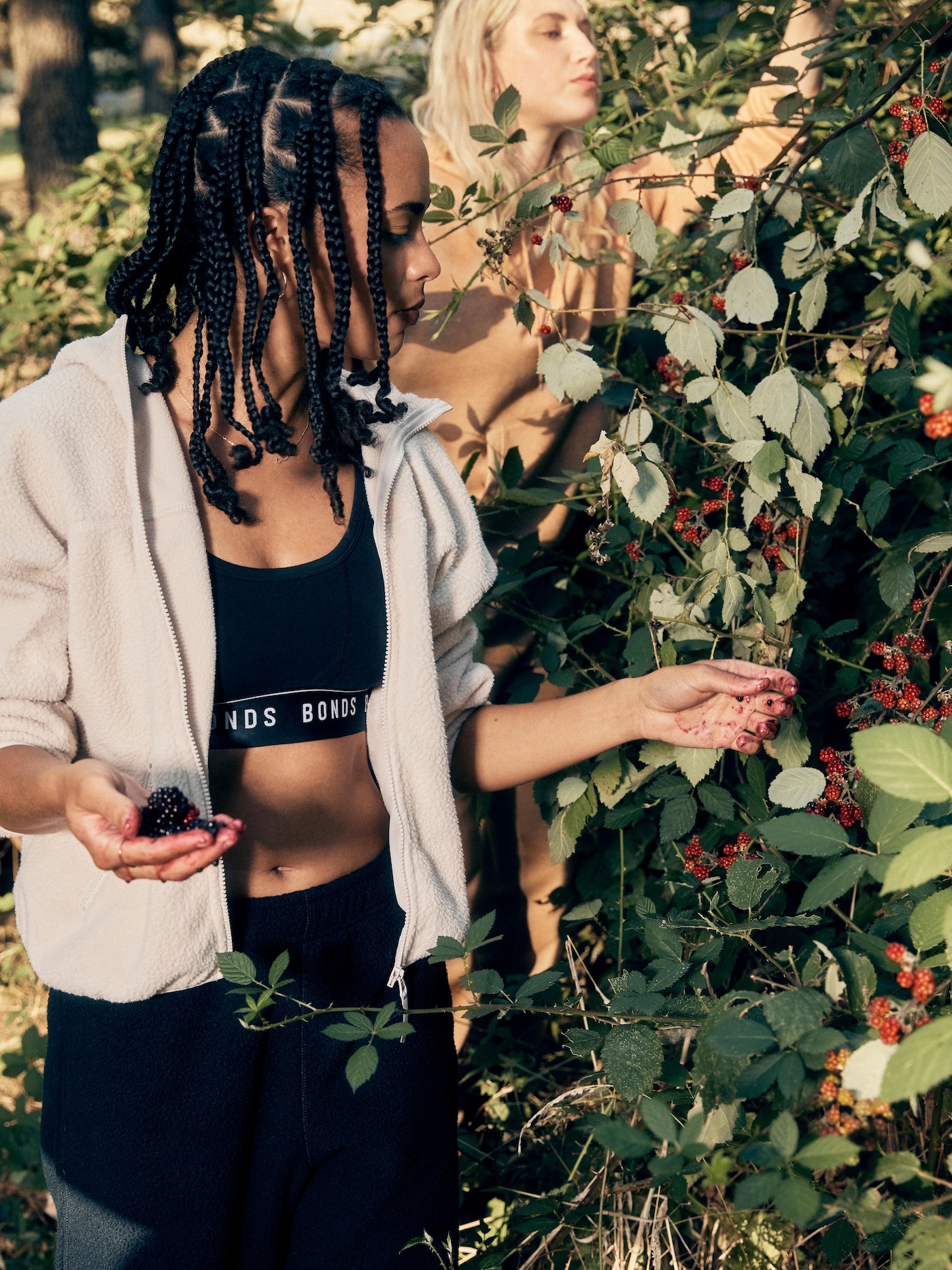 Two women in athletic wear foraging for berries in a sunlit outdoor scene with lush greenery. One woman holds dark berries.