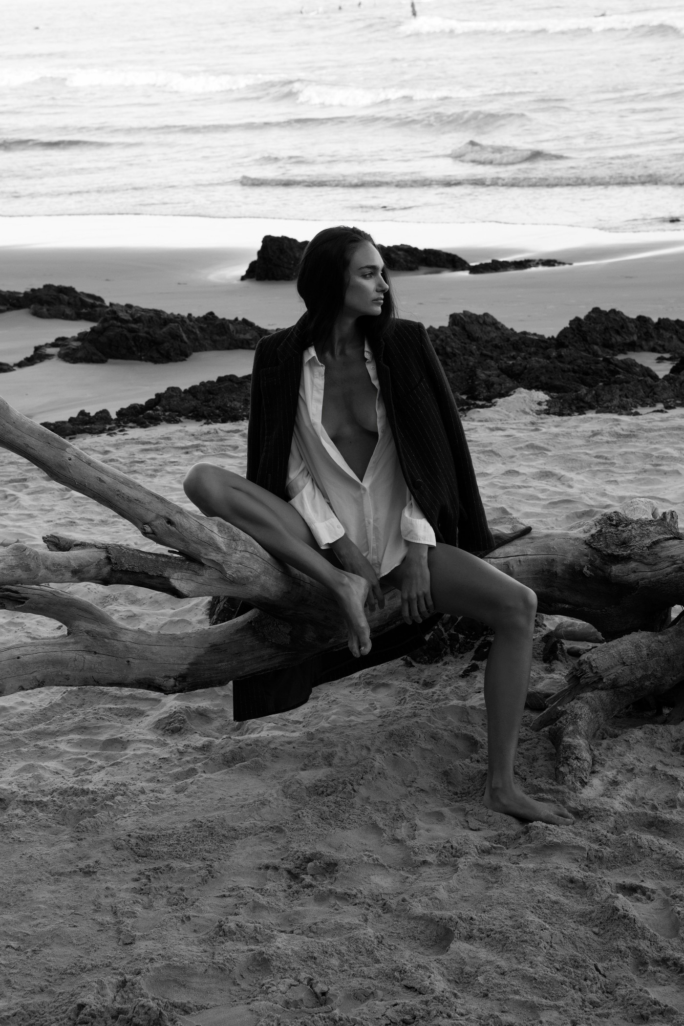 Black and white photo of a female fashion model sitting on driftwood at a beach in an unbuttoned shirt and jacket at dusk.