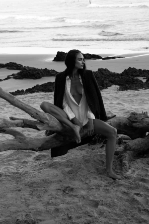 Black and white photo of a female fashion model sitting on driftwood at a beach in an unbuttoned shirt and jacket at dusk.