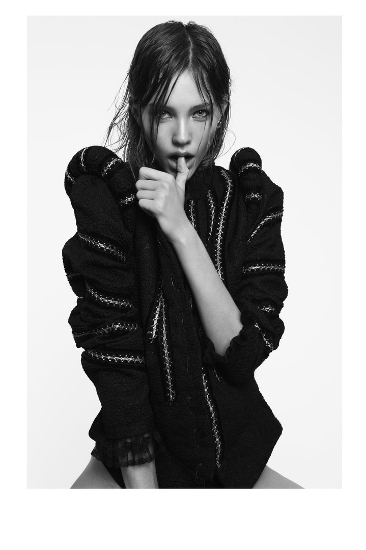 Black and white studio shot of a woman with wet hair wearing a decorative dark jacket and looking at the camera with a neutral expression.