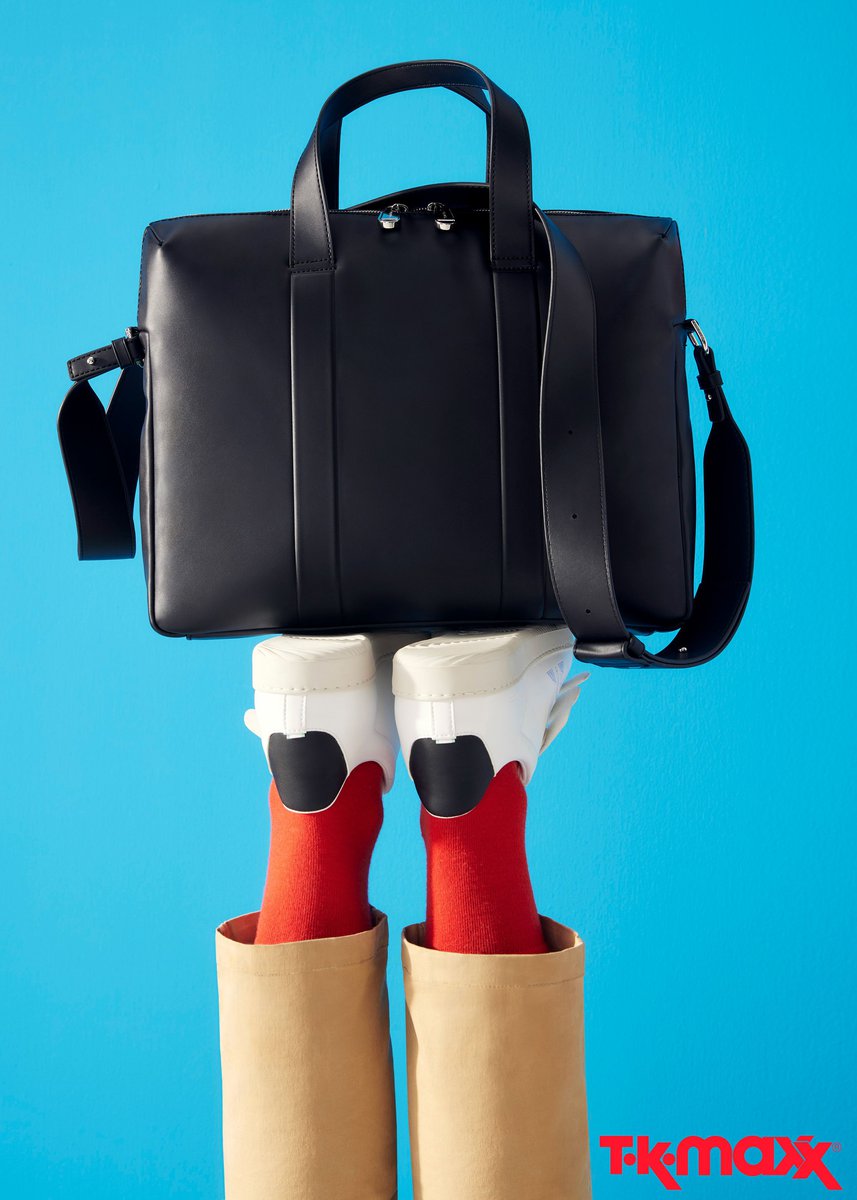 Black leather satchel bag floating over a pair of white sneakers and red socks; legs in paper bags on a vibrant blue background.