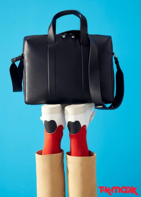 Black leather satchel bag floating over a pair of white sneakers and red socks; legs in paper bags on a vibrant blue background.